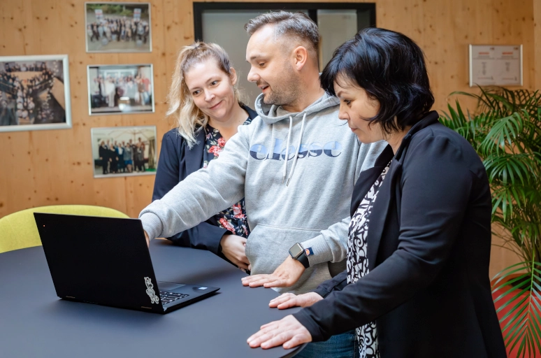 Ein Mann in einem grauen Hoodie steht an einem schwarzen Hochtisch und erklärt zwei Frauen in schwarzen Blazern das Thema Wochenendzuschlag. Dieses ist auf einem Laptop in ihrer Mitte zu sehen.
