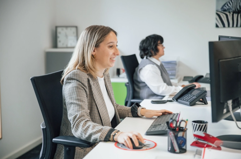 Eine Frau in einem beigem Blazer sitzt an einem weißen Tisch und arbeitet an einem Computer. Sie bereitet die Lohnfortzahlung für einen kranken Mitarbeiter vor. Im Hintergrund sitzt eine Frau in weißer Bluse und grauer Weste.