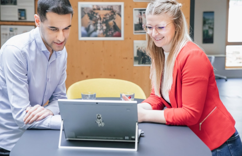 Eine Frau in einem roten Blazer steht an einem schwarzen Hochtisch. Gegenüber von ihr steht ein Mann in hellblauem Hemd. Beide schauen auf einen Laptop in ihrer Mitte und berechnen die Beitragsbemessungsgrenze.