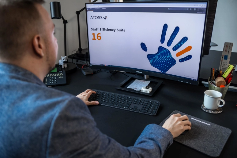 Ein Mann sitzt vor einem PC auf dem gerade ATOSS gestartet wird, um zu schauen ob der Minusstunden hat