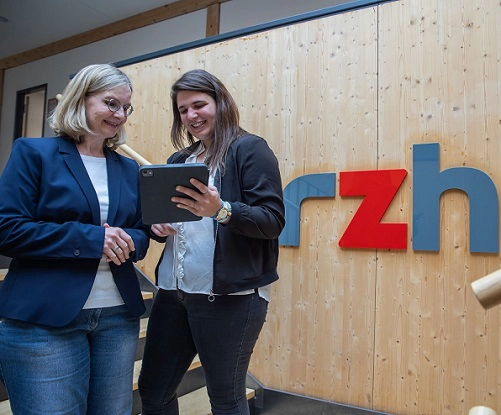 Eine braunhaarige Frau mit schwarzer Jacke, weißen Blazer und grauer Hose zeigt einer blonden Frau mit blauen Blazer, weißem T-Shirt und blauer Jeans auf einem Tablet das interne Kontrollsystem.